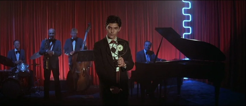 Blue Velvet (1986) movie still: fullbody — A night club stage features a jazz quartet positioned in front of heavy red curtains. A young man stands…