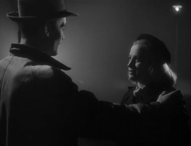 Music in Darkness (1948) movie still: medium — A dark, fog-covered exterior scene at night with a single faint light source visible. A man in a hat…