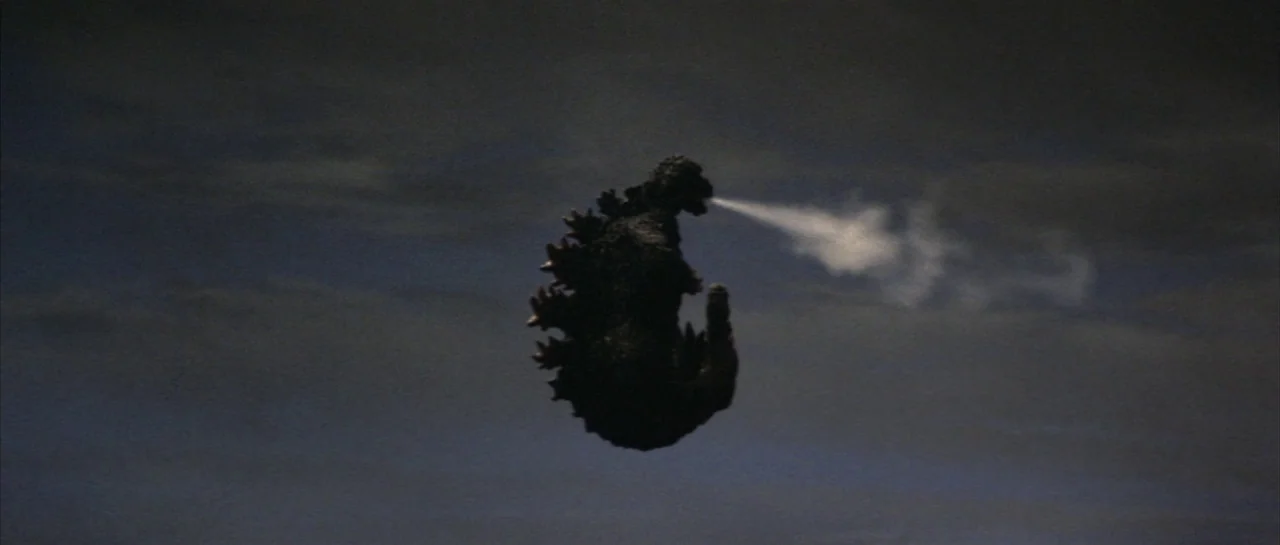 Godzilla Vs Hedorah (1971) movie still: wide — A giant lizard creature flies horizontally against a dark and cloudy night sky. The creature exhales…