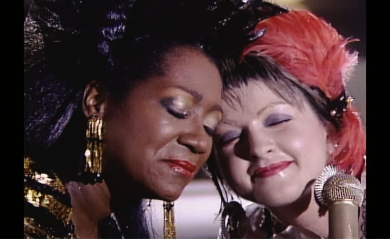 Let the Canary Sing (2023) movie still: closeup — A close-up of two women performing in a brightly lit, nondescript interior space. The women hold…
