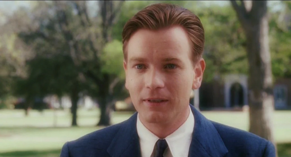 Big Fish (2003) movie still: closeup — A man stands outdoors on a green lawn with blurred trees and brick buildings. The man faces the camera while…