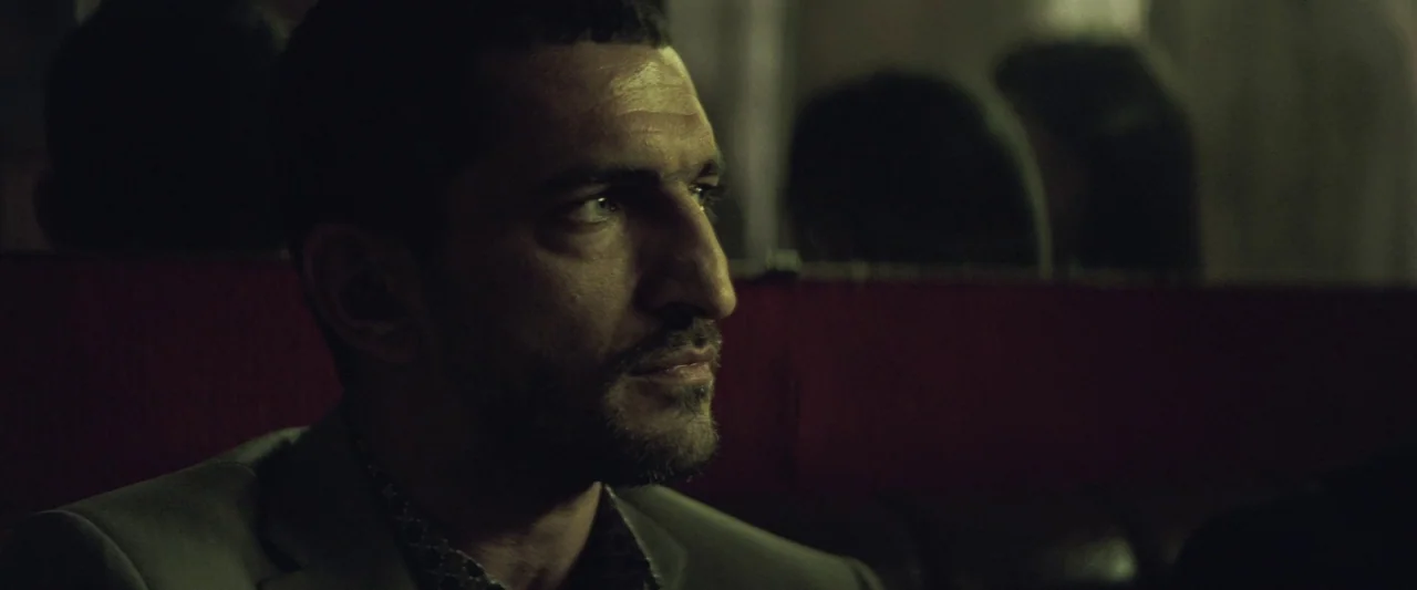 Colt 45 (2014) movie still: closeup — A dark indoor space with blurred background shapes suggesting tiered seating. A man with short dark hair sits…