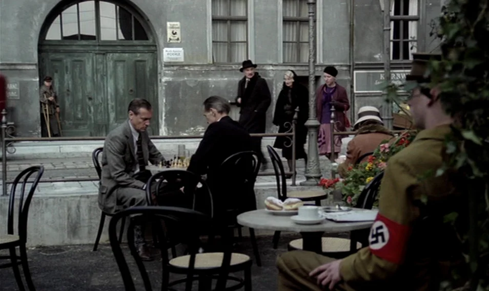 Despair (1978) movie still: wide — An outdoor cafe setting features men playing chess at tables in front of a building. A German soldier with a red…