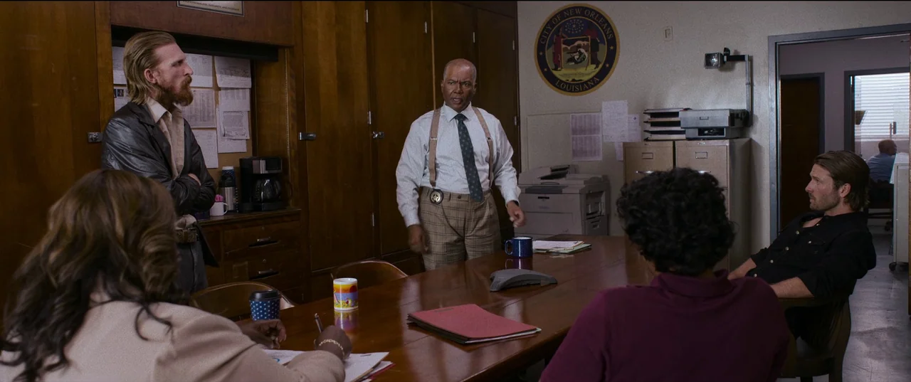 Hit Man (2023) movie still: wide — A wood-paneled room displays a City of New Orleans police seal on the wall. A senior officer stands before four…