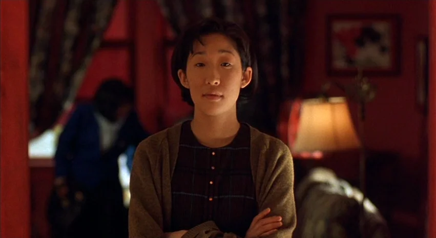 Double Happiness (1994) movie still: medium — A woman stands in a living room characterized by deep red walls and ambient lighting. A secondary…