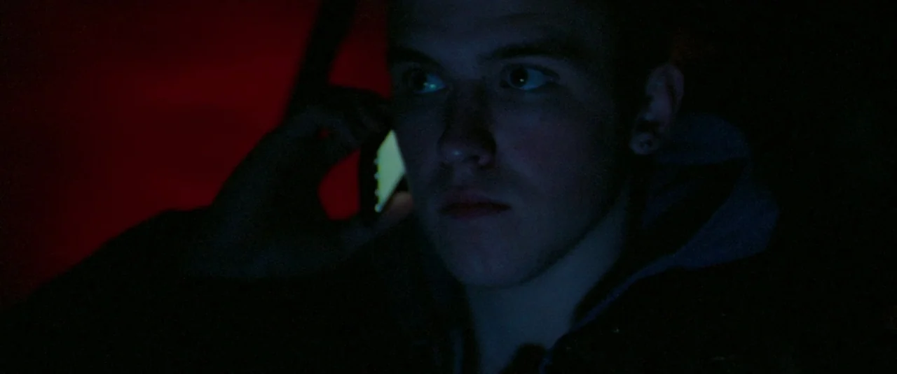 Catch Me Daddy (2014) movie still: closeup — A dark car interior is filled with shadows and sharp color contrasts. A young man sits in the dark…