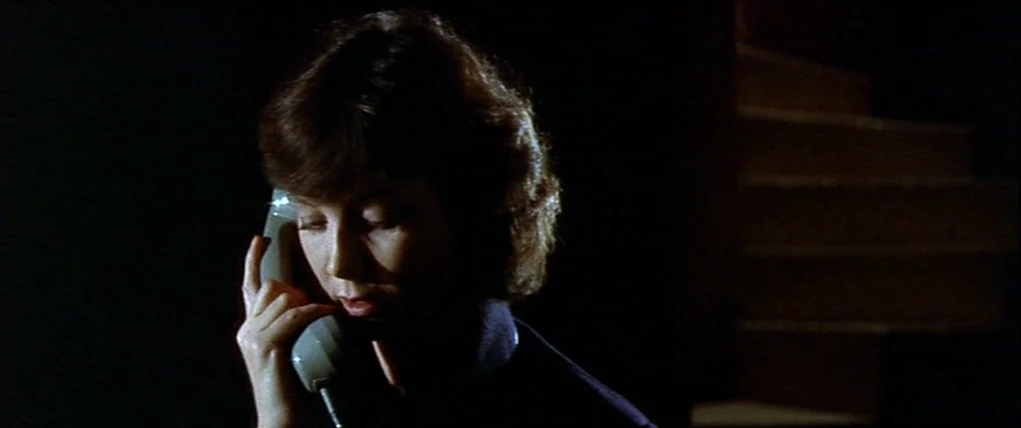 Love Unto Death (1984) movie still: closeup — A woman stands near a dark wooden staircase at night. She listens to a beige telephone handset held…