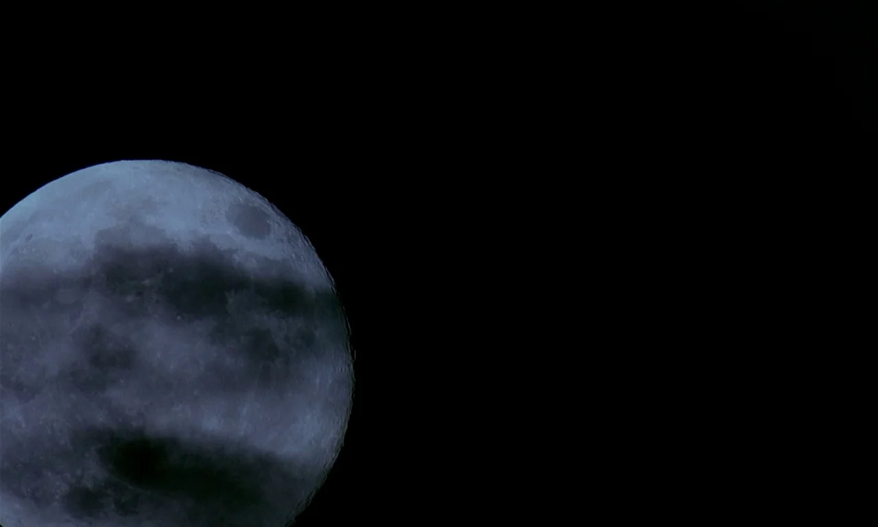 Microcosmos (1996) movie still: wide — A partial view of the moon sits against a vast expanse of empty space. Dark clouds pass horizontally across…
