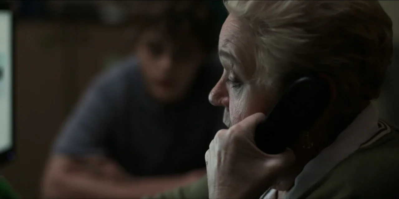 Bring Her Back (2025) movie still: closeup — A dimly lit room features an older woman in profile talking on a phone. In the out-of-focus background,…