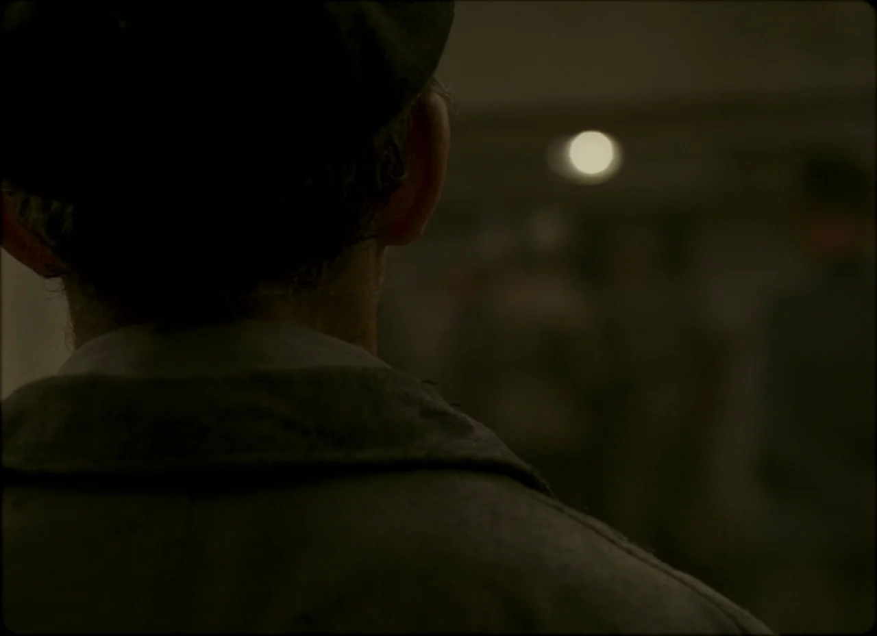 Son of Saul (2015) movie still: closeup — A dark, blurred corridor is visible behind a person standing in the foreground. The subject looks ahead at…