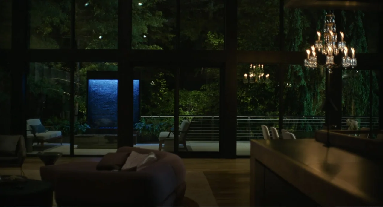 Turtles All the Way Down (2024) movie still: establishing — A dark modern living room overlooks an outdoor terrace at night through floor-to-ceiling…