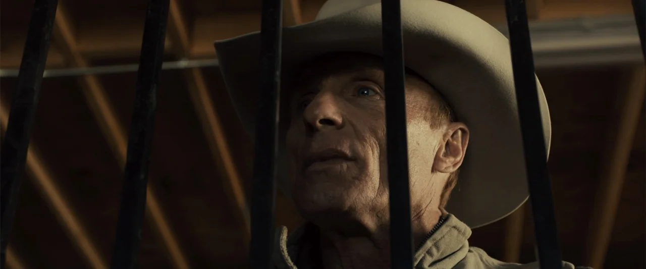 Frontera (2014) movie still: closeup — A dark indoor space with wooden rafters visible above and vertical metal bars foreground. An elderly man in a…