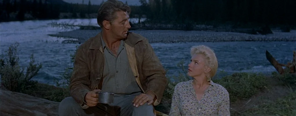 River of No Return (1954) movie still: medium — A man and a woman sit together by a flowing river at twilight. The environment includes a log,…