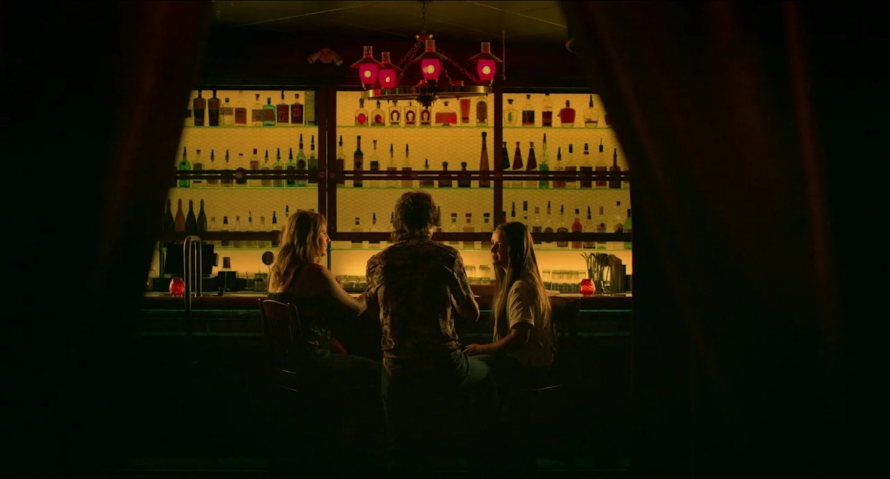 Trim Season (2023) movie still: medium — A dimly lit room with rows of backlit glass bottles lining the wall shelves. Three people sit on stools at…