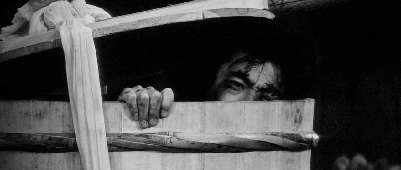 Yojimbo (1961) movie still: closeup — A man hides inside a large cylindrical wooden barrel bound with twisted bamboo strips. His hand grips the top…