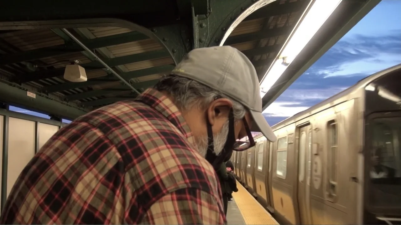 Covid Diaries NYC (2021) movie still: medium — A modern outdoor subway platform features metal structural pillars and incoming train cars. A man in…