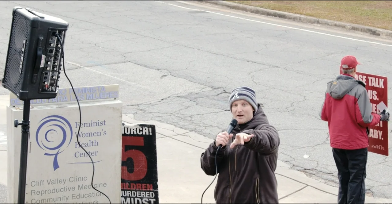 The Devil Is Busy (2024) movie still: medium — A man stands on a sidewalk in front of a medical center entrance. The man holds a microphone and…