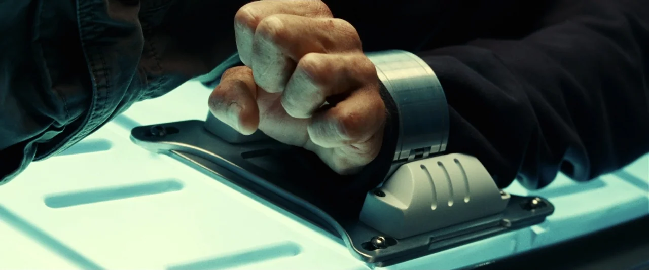 Green Lantern (2011) movie still: closeup — A close-up of a hand restrained on a glowing, white plastic and metal surface. The wrist is secured…