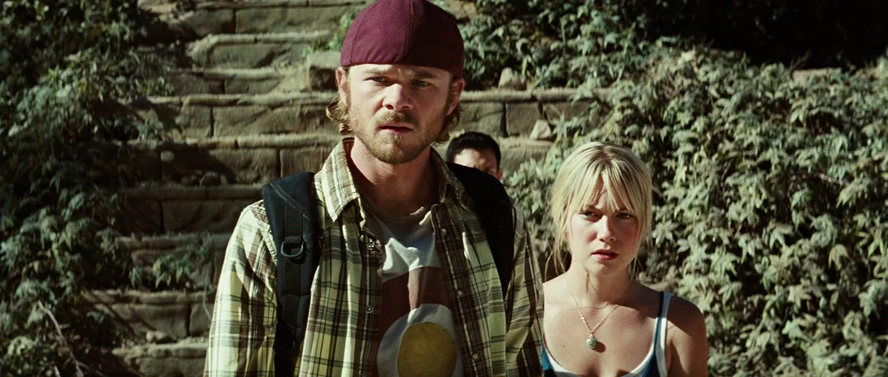 The Ruins (2008) movie still: medium — A man and woman stand on weathered stone steps covered in overgrown vines. They look ahead with serious…