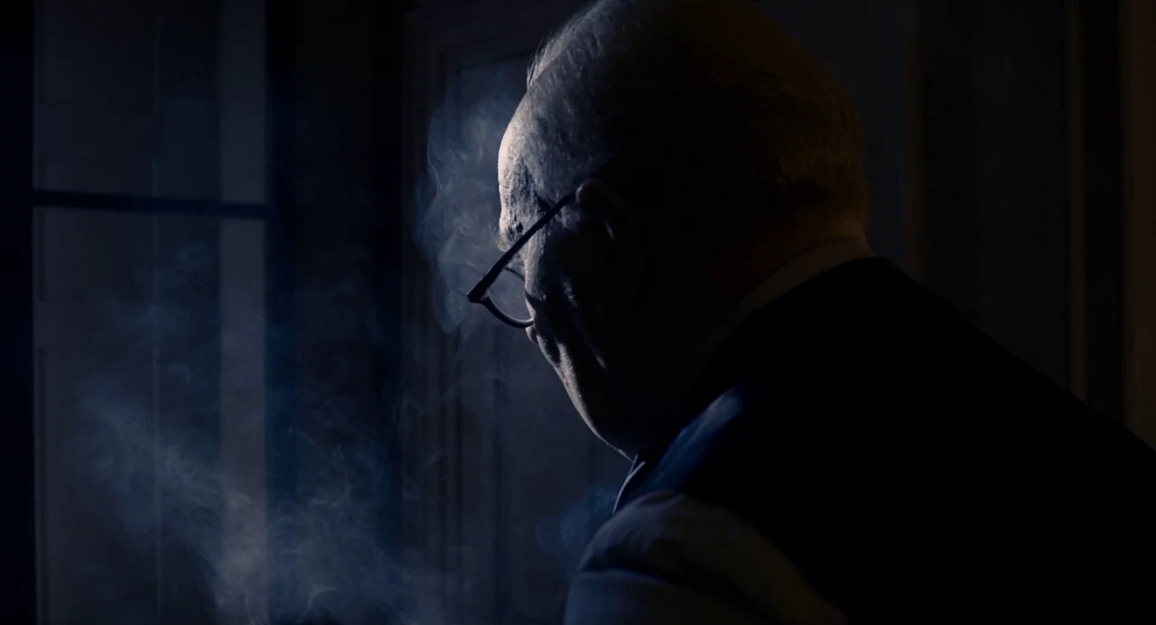 Darkest Hour (2017) movie still: closeup — An elderly man in a suit sits in a dimly lit, shadowed room. He looks down while smoke drifts past his…
