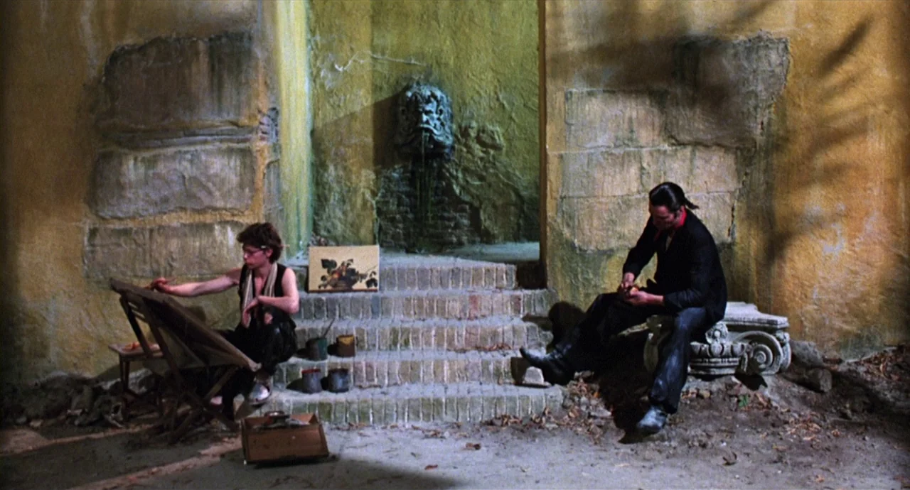 Caravaggio (1986) movie still: wide — An outdoor courtyard features yellow stone walls and a stone fountain mask on steps. A young painter works at…