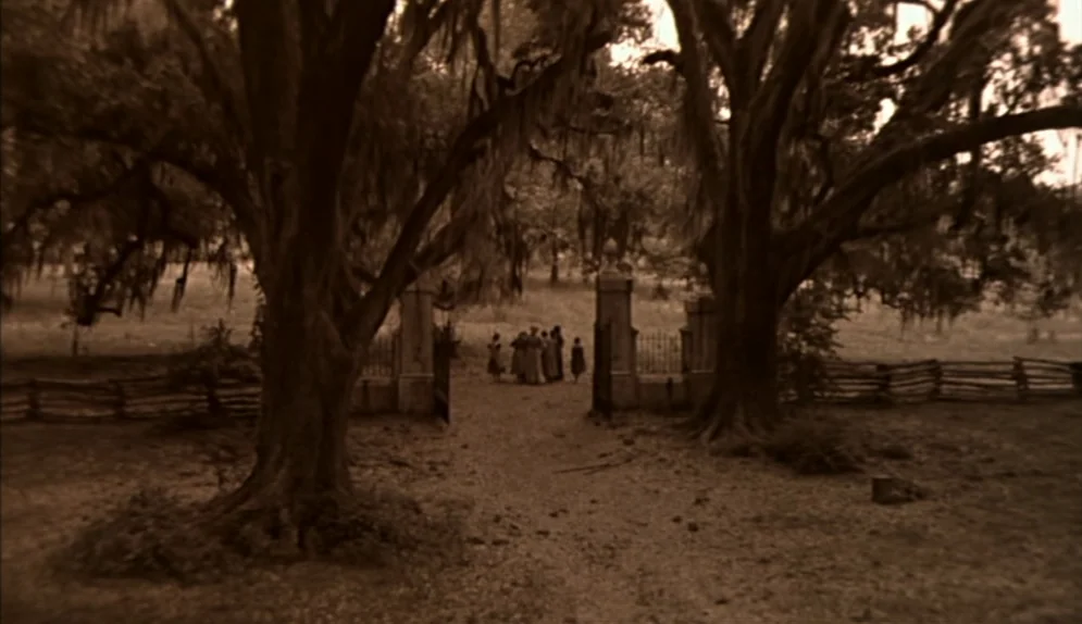 The Beguiled (1971) movie still: wide — Large oak trees with Spanish moss frame a rustic wooden fence and iron gate. A group of figures in period…