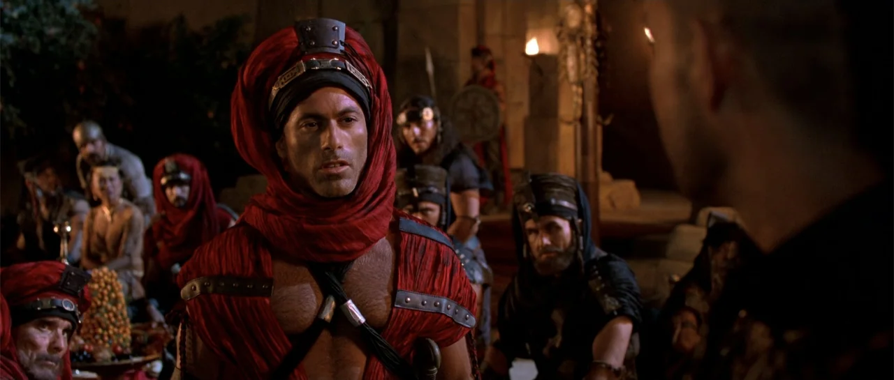 The Scorpion King (2002) movie still: medium — A dimly lit ancient stone hall is filled with people at a feast. A man in red clothing stands at the…