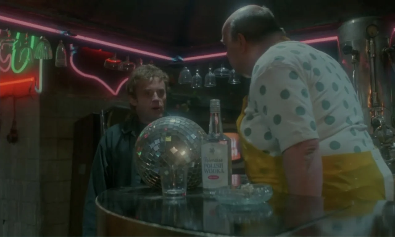 Ga-ga: Glory to the Heroes (1986) movie still: medium — A dimly lit bar interior features a glass counter with a disco ball and vodka. A younger man…
