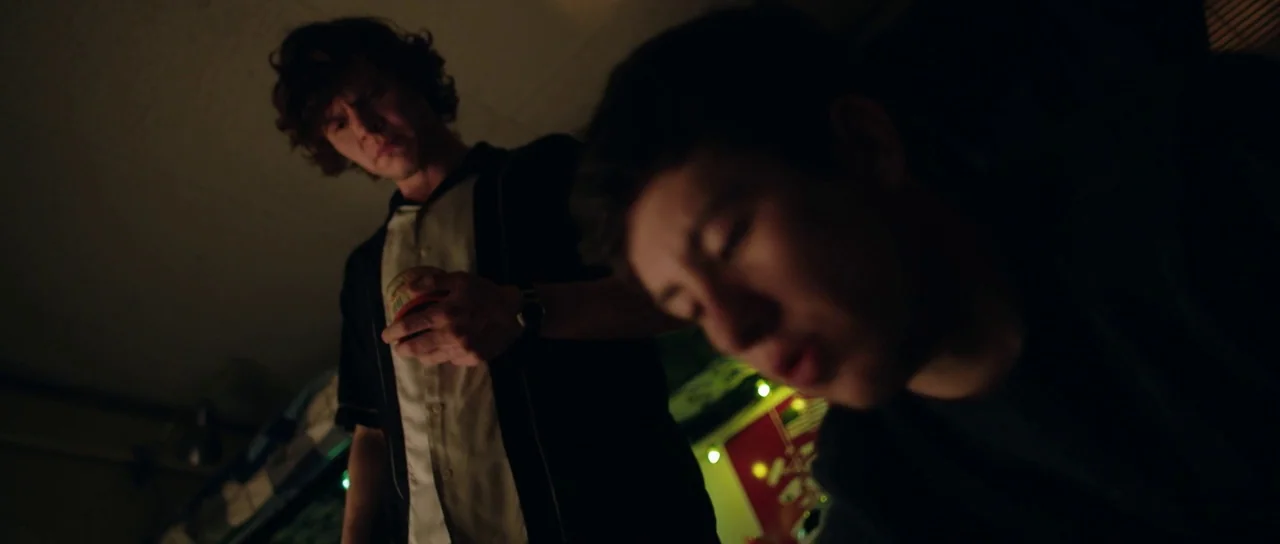 American Animals (2018) movie still: medium — A dark, dimly lit room with one person standing over another. The standing man watches the other man…