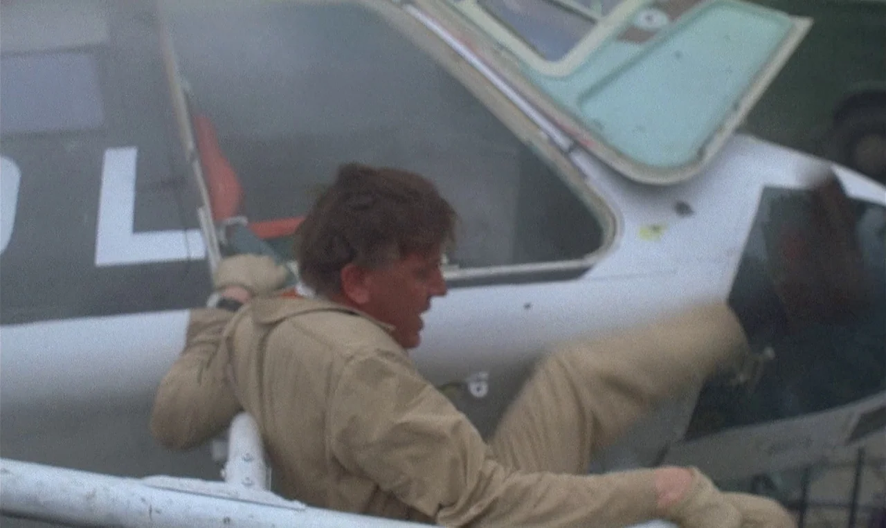Man on the Roof (1976) movie still: medium — A man sits inside the cockpit of a white and black light aircraft. The cockpit canopy is open while the…