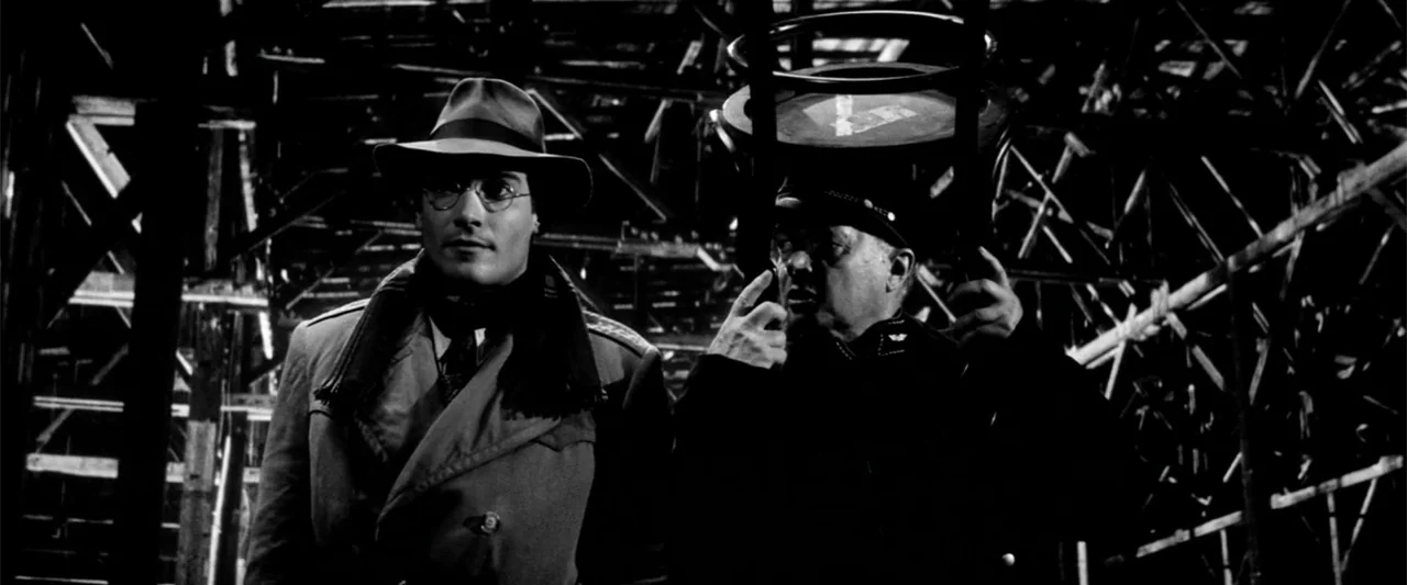 Europa (1991) movie still: medium — A complex wooden scaffolding structure frames two men in a low-light indoor setting. A man wearing a fedora…