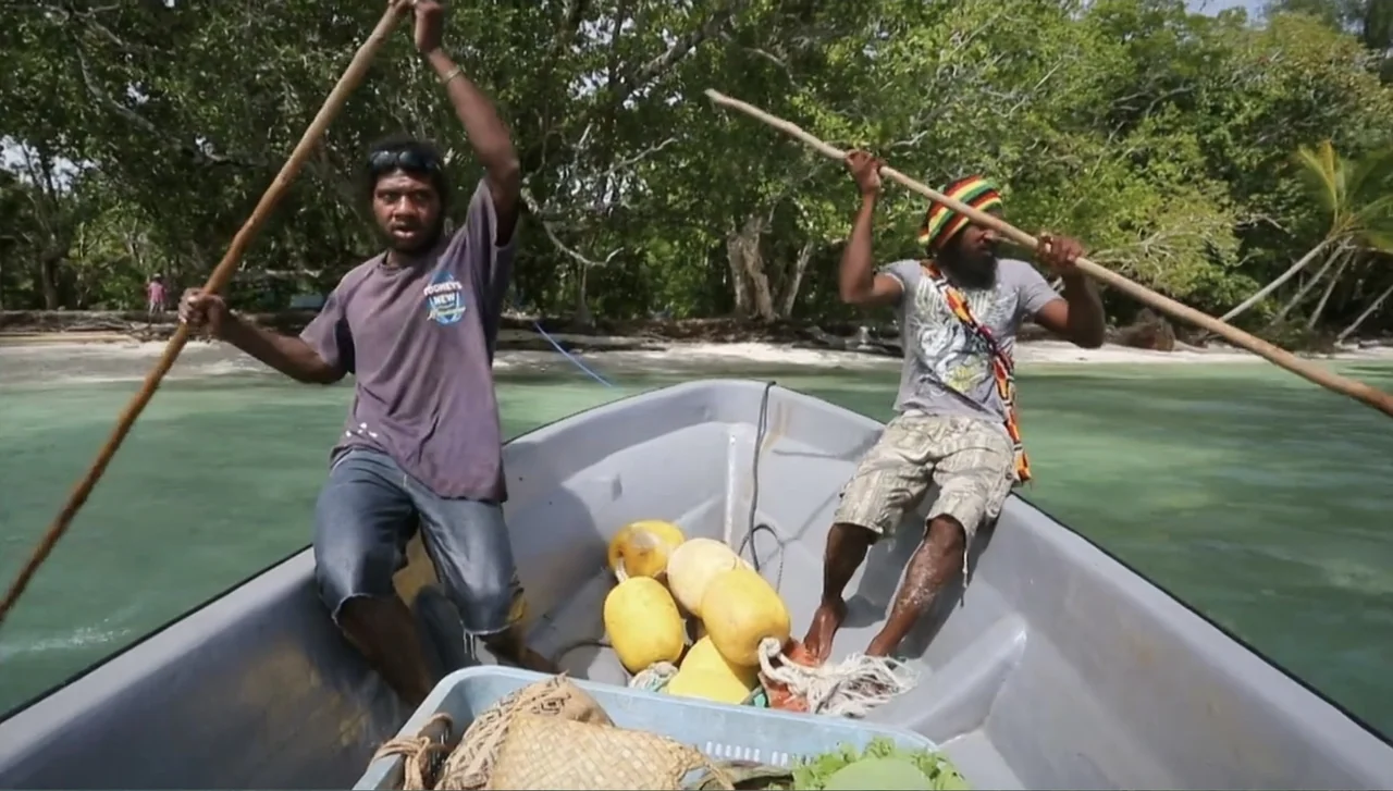 Islands of Creation (2015) movie still: medium — Two men maneuver a small plastic boat through shallow tropical waters near a shoreline. Tropical…