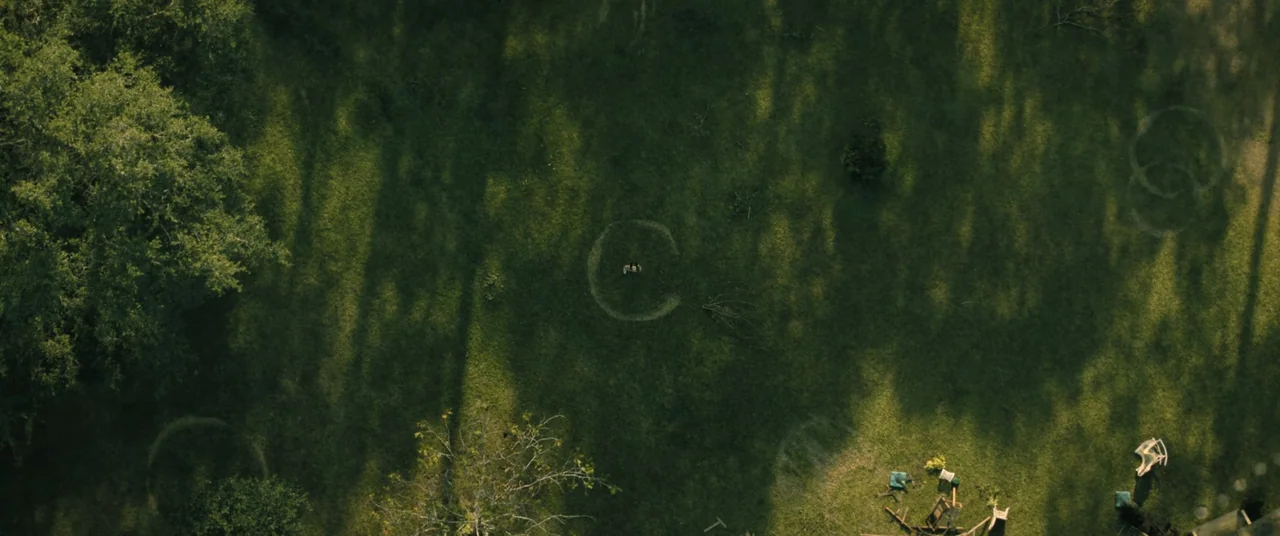 No One Will Save You (2023) movie still: establishing — An open grassy field contains several large circular markings drawn into the turf. Long tree…