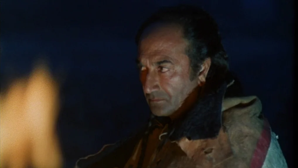 The Scarlet Letter (1973) movie still: closeup — A man sits outside at night near a small, flickering fire. He gazes off-frame into the darkness…