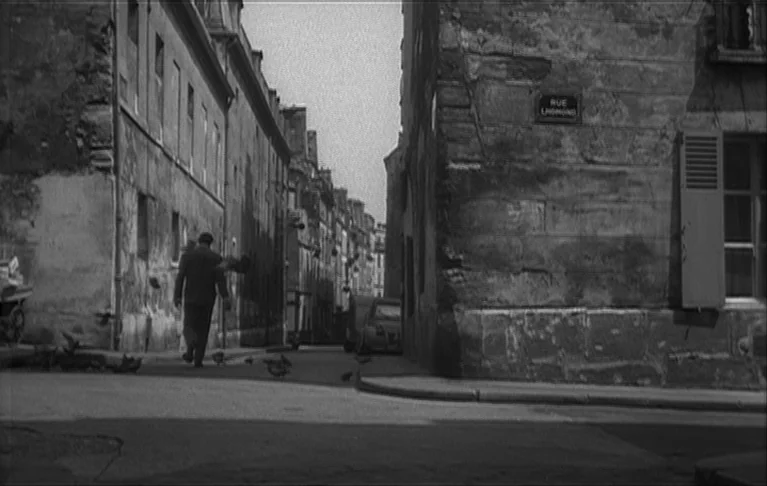 Le Signe du Lion (1962) movie still: wide — A narrow stone-walled urban passage is marked by an old French street sign. A man in a dark suit walks…