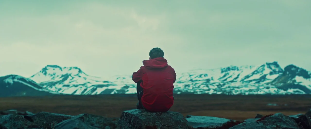 Rift (2017) movie still: wide — A vast, open plain lies before a range of snow-capped mountains under a clouded sky. A lone person sits on a rock,…