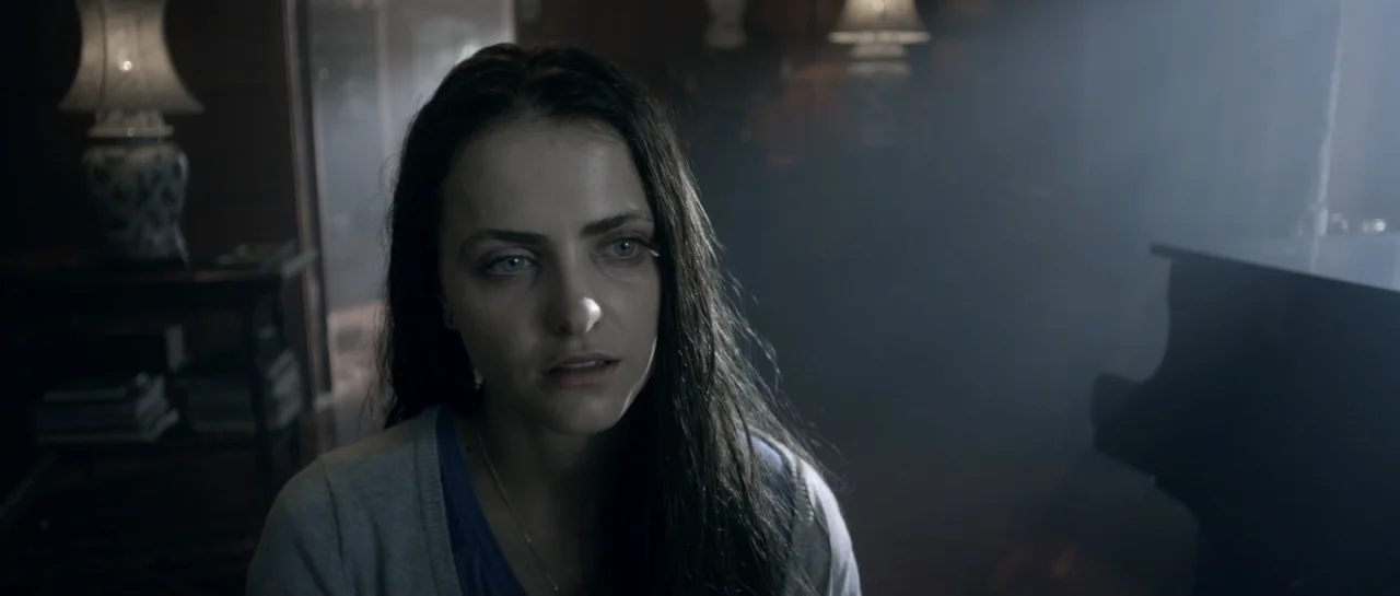 The Lullaby (2019) movie still: closeup — A dark living room filled with ambient smoke and dimly lit by a table lamp. A woman with long dark hair…