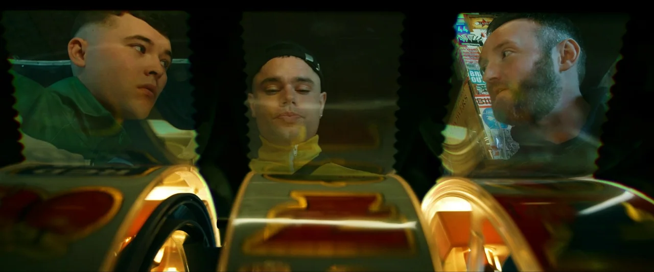 Kneecap (2024) movie still: medium — An interior space features three men leaning over a glowing, mechanical slot machine display. The low-angle…