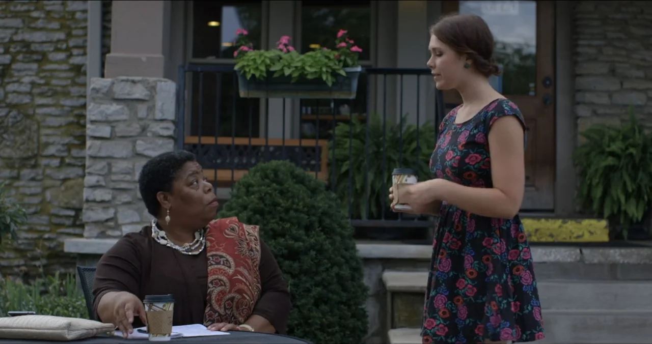 Faith, Love & Chocolate (2018) movie still: medium — An outdoor seating area is positioned in front of a stone building with planters. Two women…