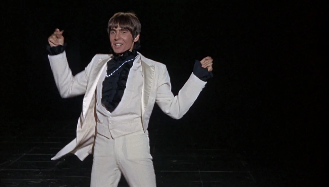 Head (1968) movie still: fullbody — A dark, empty void serves as a minimalist stage backdrop. A man in a bright white tuxedo suit stands centered…