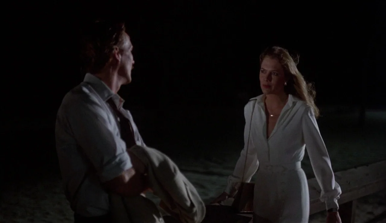 Body Heat (1981) movie still: medium — A dark beach at night with sand visible in the foreground and a wooden rail. A man and a woman stand facing…