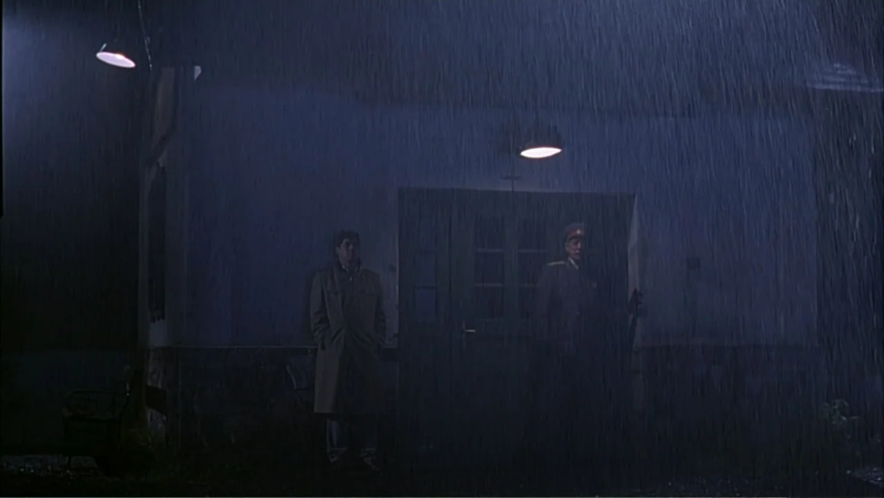 Citizen X (1995) movie still: wide — A plain building exterior is illuminated by two overhead lamps during a dark rainstorm. A man in a trench coat…