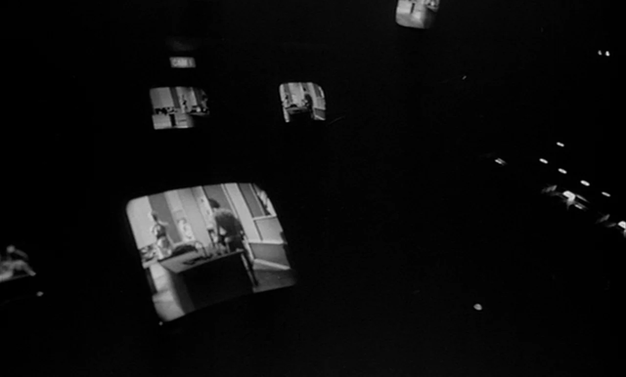 Stereo (1969) movie still: wide — A dark television control room features multiple monitors displaying grayscale footage of internal sets. Each…