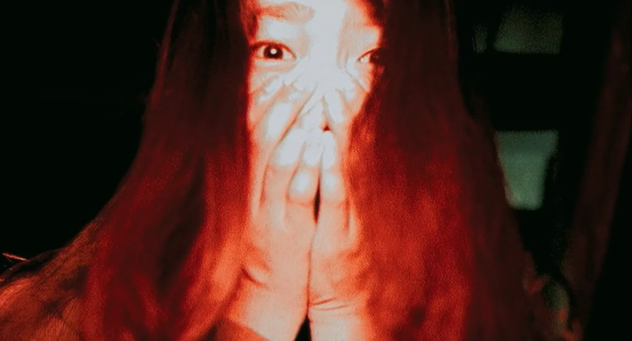 Uzumaki (2000) movie still: closeup — A person stands in a completely dark space with only their face illuminated. The subject holds their hands…