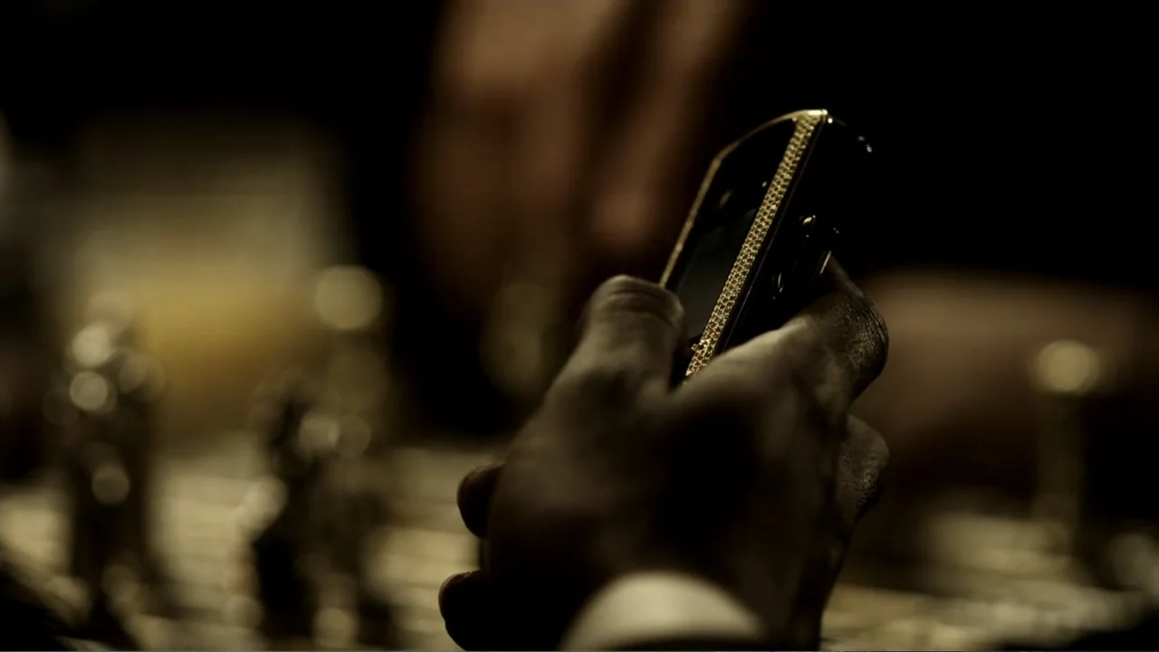 Assassination Games (2011) movie still: closeup — A dark interior room features a chess set blurred in the foreground. A hand holds a high-end black…