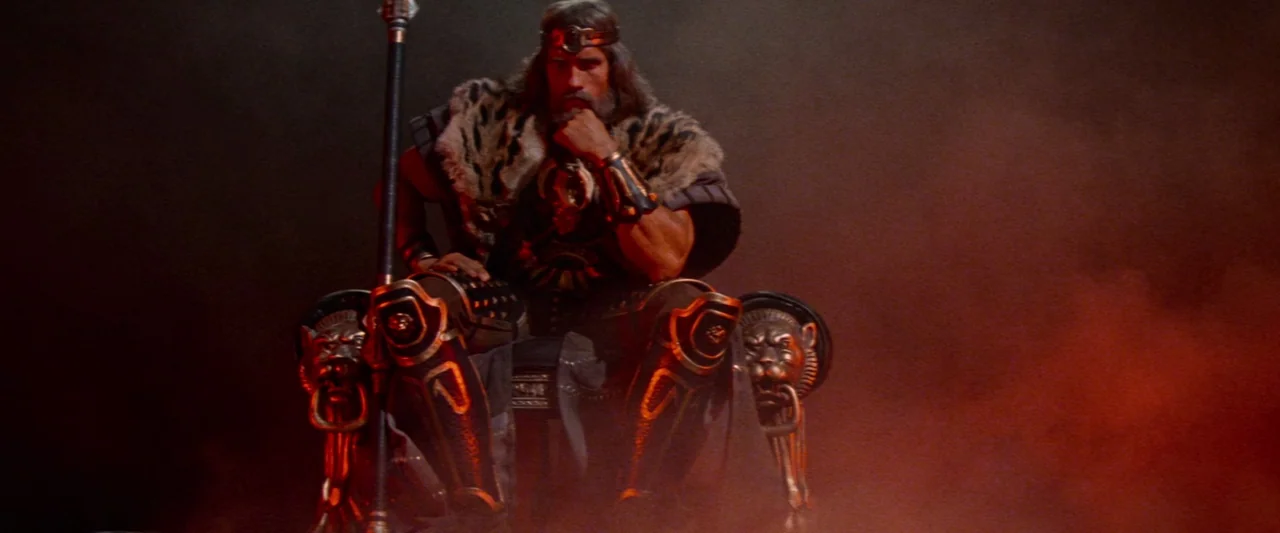 Conan The Barbarian (1982) movie still: medium — A dark, smoky space featuring an ornate throne with sculpted lion heads on the armrests. A man in…