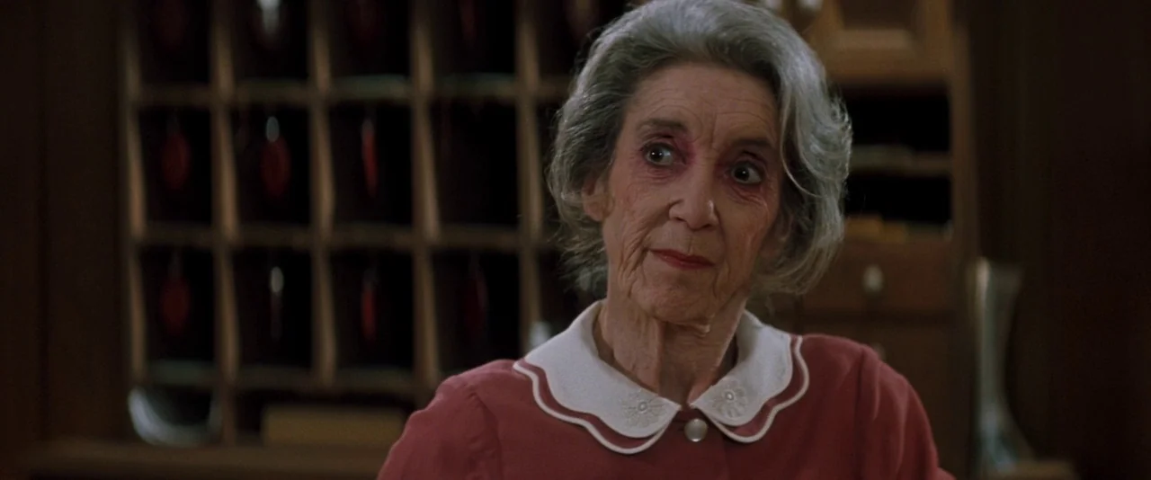 In The Mouth of Madness (1994) movie still: medium — The background features a wooden grid wall used for sorting mail in a hotel. An elderly woman…