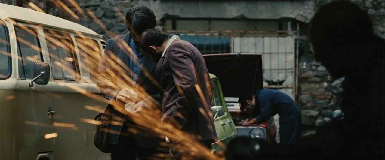 Argo (2012) movie still: medium — A cluttered outdoor auto repair yard with vintage cars and a stone wall background. Two men in formal jackets…