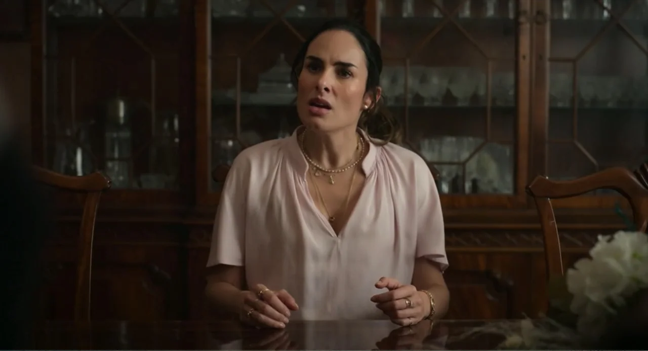La posada (2023) movie still: medium — A dark wood dining room with a glass-fronted china cabinet in the background. A woman sits at a table while…