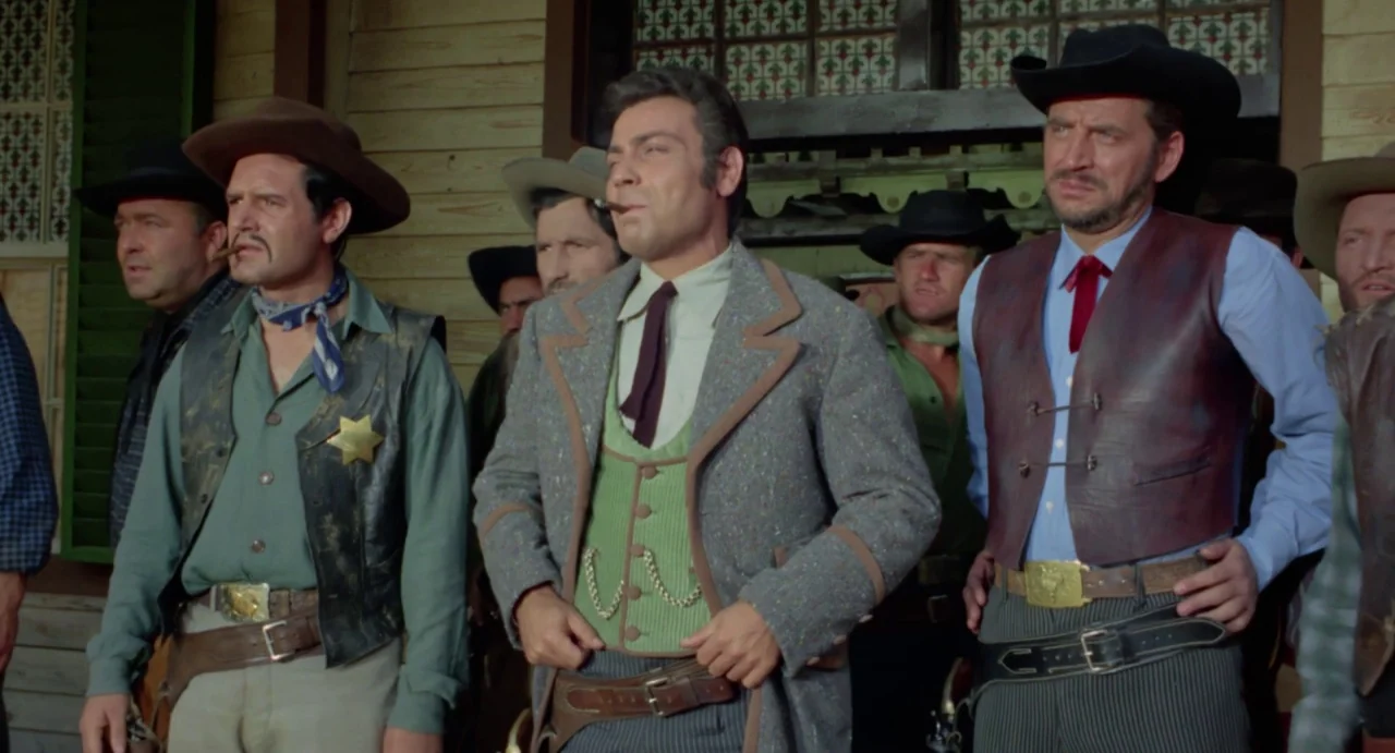 Johnny Colt (1966) movie still: medium — A group of men in historical Western clothing stands on a rustic wooden porch. Several men with guns and…